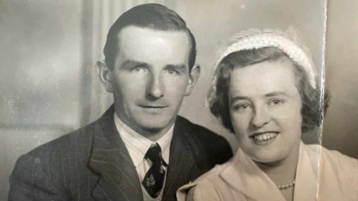 Old black and white photograph of a couple smiling.  The man is dressed in a jacket and tie and has slicked back hair.  The lady is wearing a small hat, and a light coloured blouse and a pearl necklace.