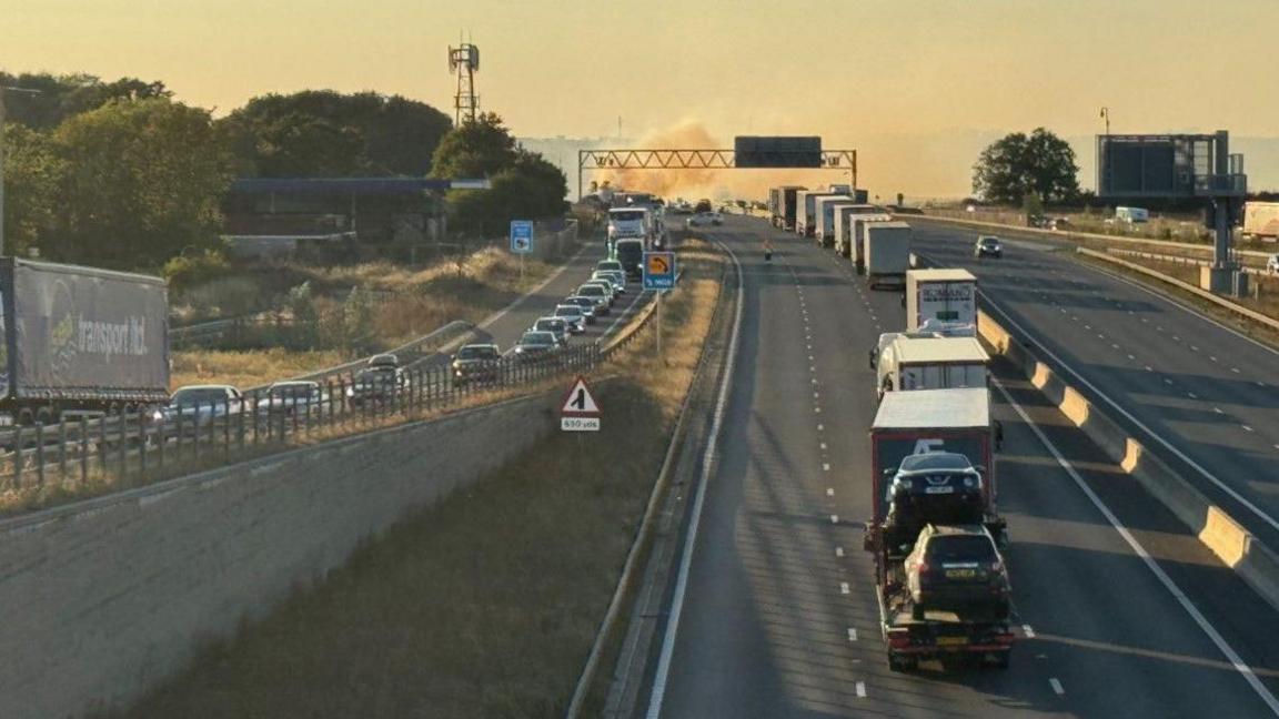 Cars and a lorry are driving up a bridge next to a three-lane road. There is a line of lorries waiting for directions and smoke can be seen in the distance