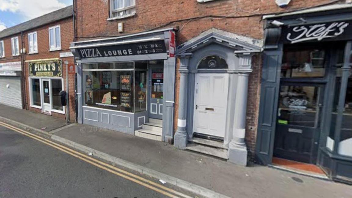 A Google Streetview shot of a street with two-storey buildings, which have small shops and restaurants on the ground floor. One shopfront has a black sign that says "Pizza Lounge" in grey letters.