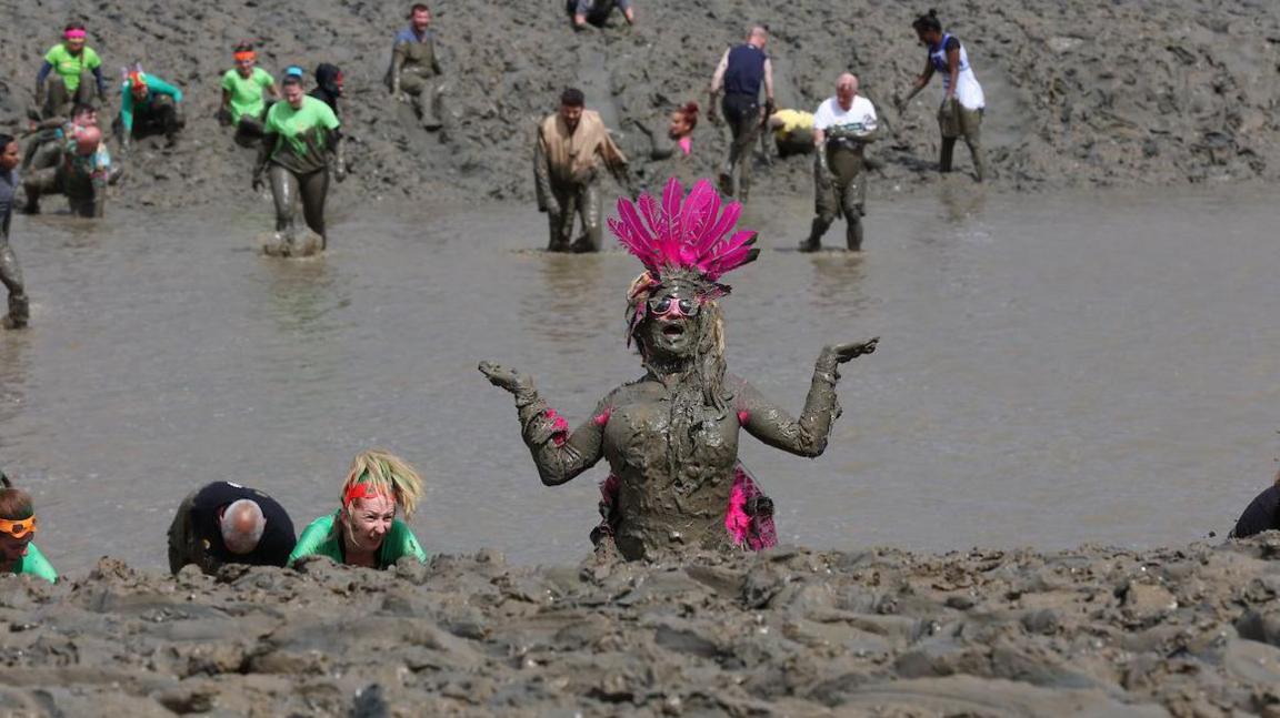 A woman with a pink-feathered head-dress is visible over the bank of the estuary. She has her arms bent at the elbows and outstretched at her sides with her palms facing upwards. A stretch of water is visible between the mud in the foreground and the bank in the background. There are about 20 other mud racers visible.
