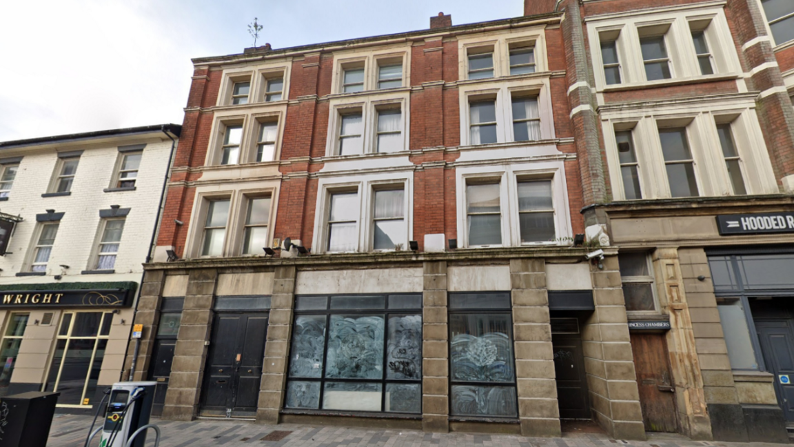 The empty building has adjoining buildings either side. It has four floors and the downstairs windows are smudged and smeared. There are windows above on each floor.