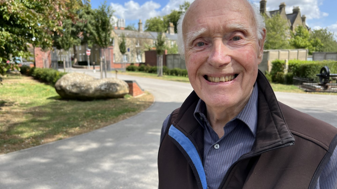 Jack Hayward smiles in a park with the railway village behind him and the DMJ tower in the distance