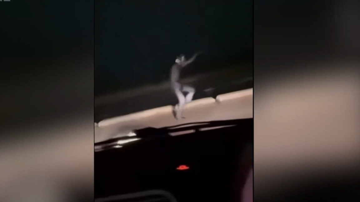A picture taken from the inside of a car which spots a man running in front of the car in a black cat suit. His arms are raised and is running fast. The headlights of the car illuminate the man in the dark night.