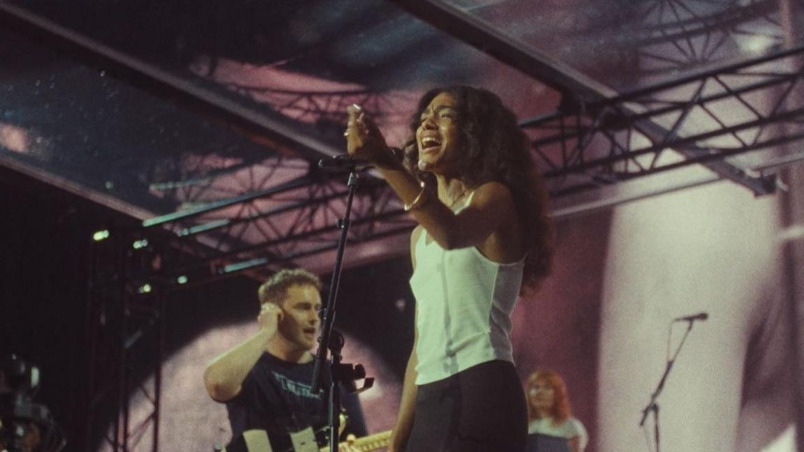 Sam Fender appears in the background holding onto his ear, wearing a black T-Shirt with his guitar also visible. Olivia Dean is at the front singing into a microphone wearing a white tank top.