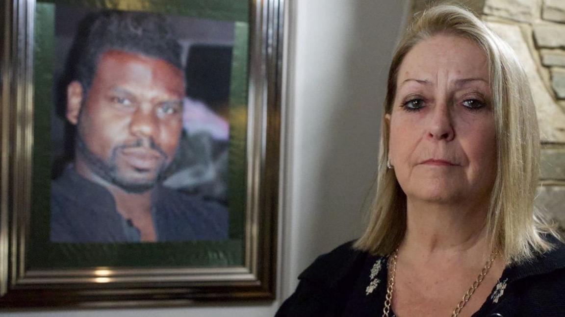 Carole Charles, a middle-aged woman looking sad. A photo of her husband is visible on the wall behind her. She is made up smartly and wears a black top and a gold-coloured chain. She has blonde hair coming down to her shoulders.