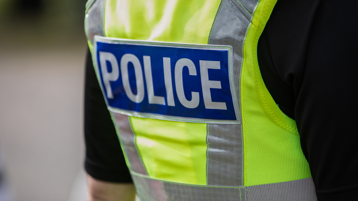 The back of a police officer's back, showing his green hi-vis jacket which says police on it in a blue box with white text