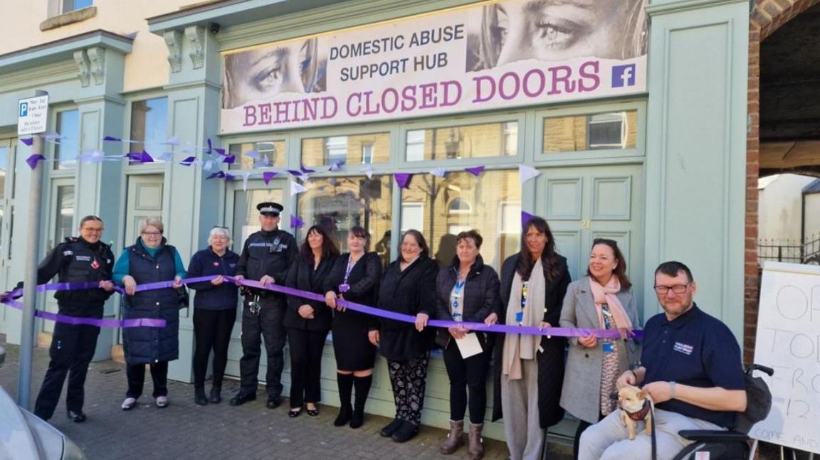 Police officers and other people involved standing behind a purple ribbon in front of the Behind Closed Doors centre in Fleetwood