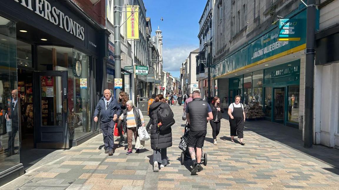 People with shopping bags walk through Douglas City Centre. The shops has bright signage, with their names in bold writing.