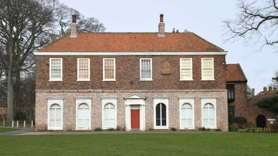 A large brick-built Georgian building, with a red tile roof and red front door. There is a lawn to the front of the building.