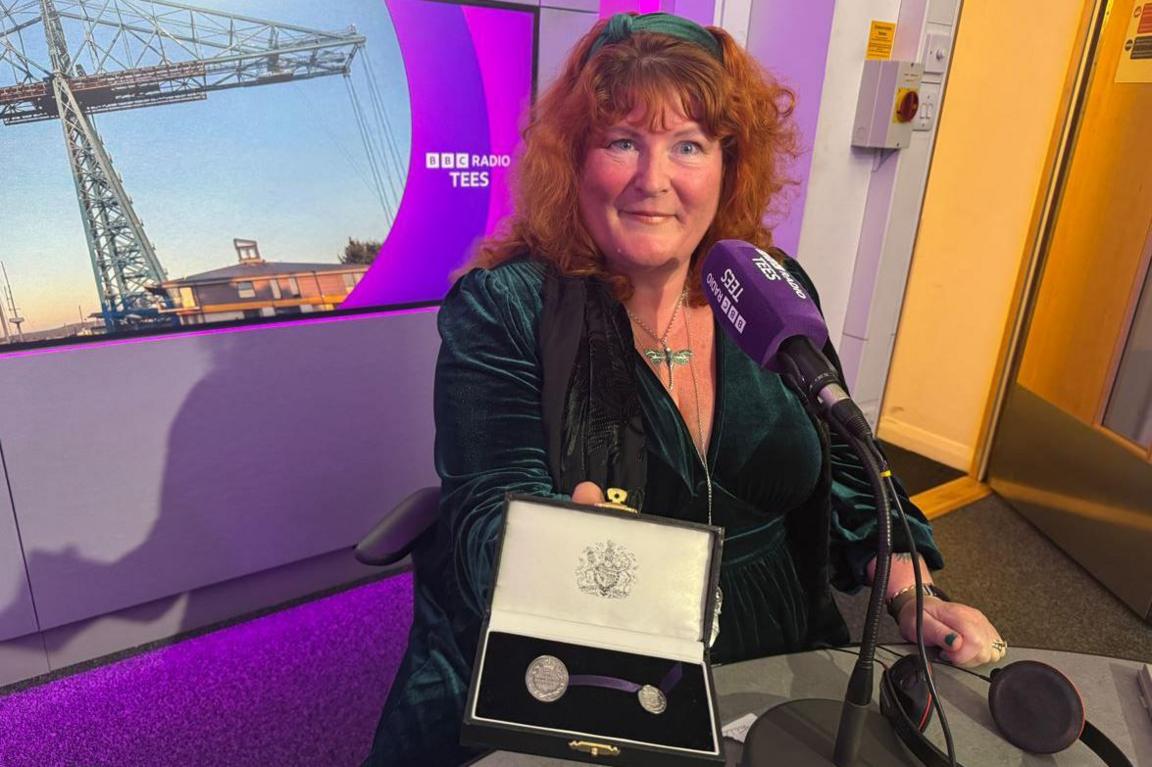 Rebecca Lombard-Earl is sitting in the purple BBC Radio Tees studio. She is holding up the Elizabeth Emblem to the camera, which is a velvet box with silver medals nestled inside. Ms Lombard-Earl, who has ginger hair and is wearing a green dress, is smiling at the camera.