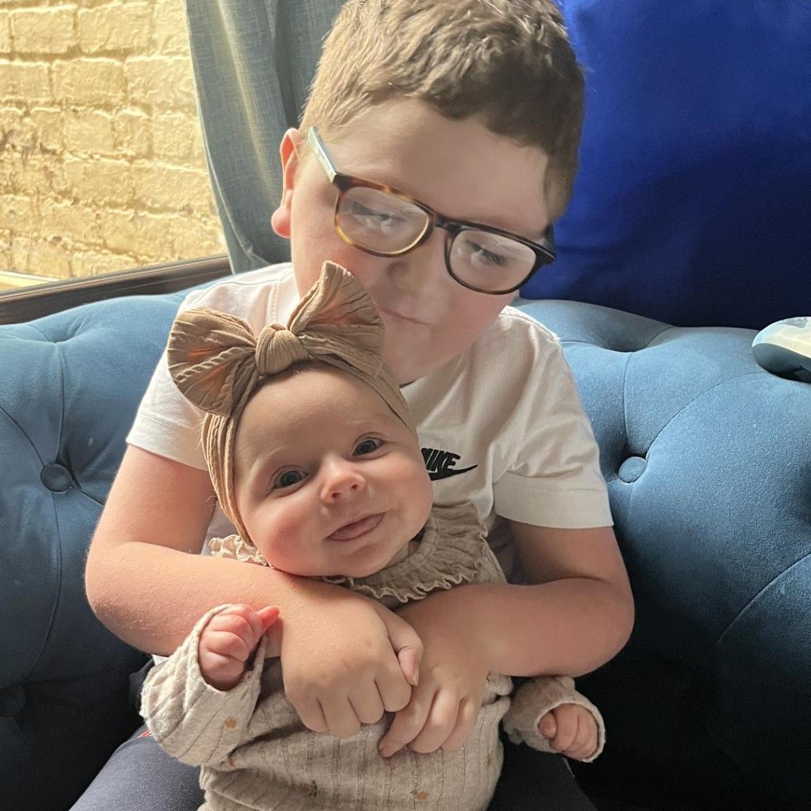 A five-year-old boy sits on a blue sofa and holds his baby sister, who is sitting in his arms. The boy has short brown hair and wears dark-framed glasses and a white T-shirt. His sister wears a cream-coloured jumper and matching headband with a large bow. She is smiling.