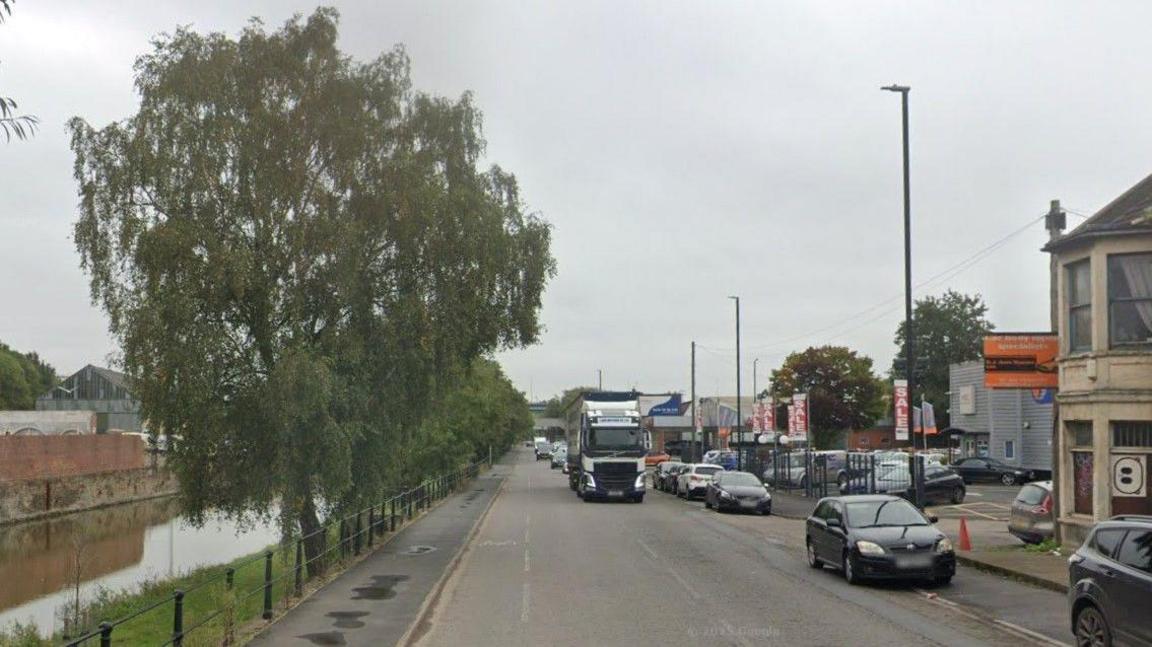 Feeder Road in Bristol with lorries and cars travelling down. It is next to a canal and has a grey sky in the background.