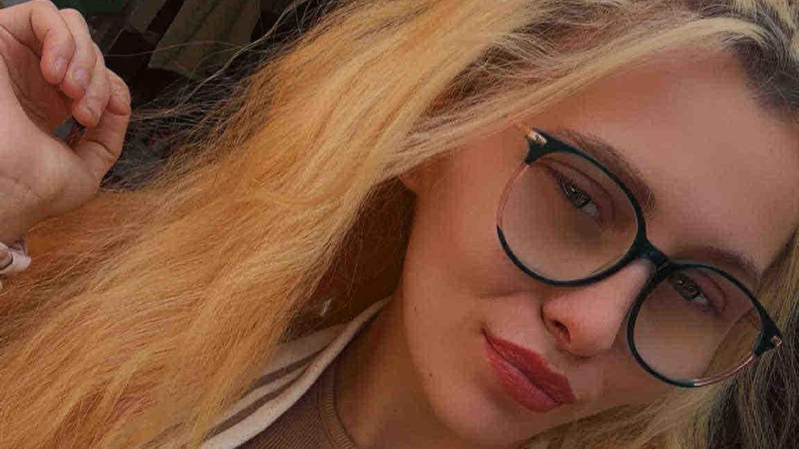 A young woman with glasses has long, wavy hair dyed blonde. One hand is raised near her head, and the background shows a dimly lit room.