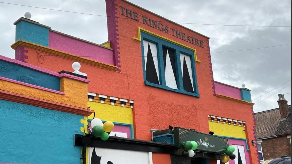 A building painted orange, blue. pink, yellow and with black and white accents. The words "The Kings Theatre" are printed at the top. Balloons are attached to the door.