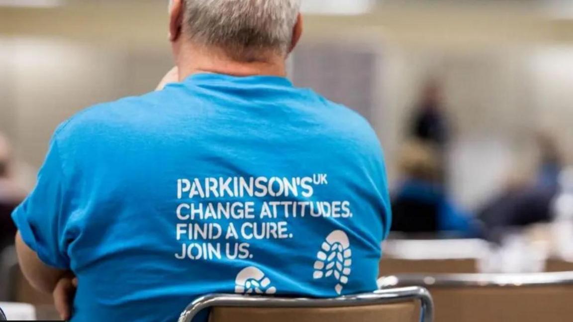 Middle-aged man with grey hair from behind wearing a blue Parkinson's UK t-shirt with the logo "Change attitudes. Find a cure. Join us"