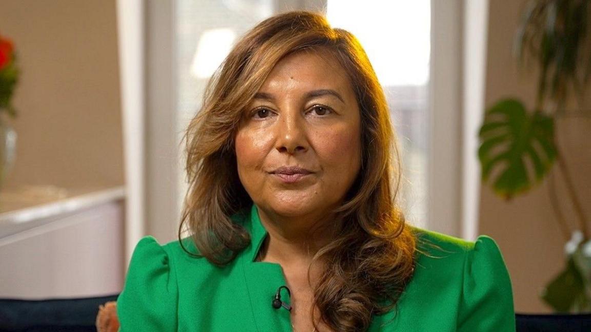 Ani Naqvi is seen from shoulders up with long brown hair wearing a green dress looks directly at the camera, seated in a brightly lit space.