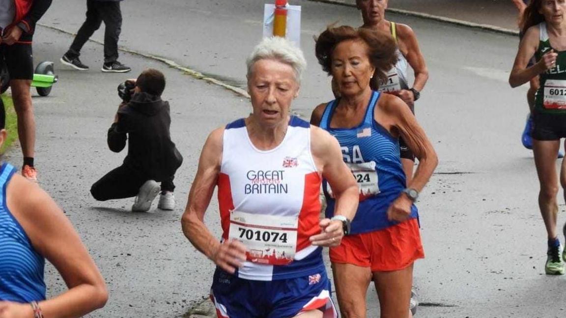 Dot Kesterton wearing the red, white and blue running vest of Great Britain, competing in the 2022 World Masters Athletics Championships in Tampere, Finland. She's surrounded by other female runners in the race and is running along a closed road.