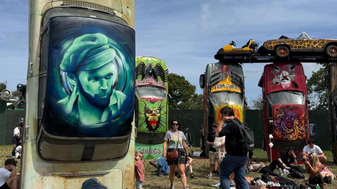 Portrait of Michael Eavis from the 1970s on the roof of a car which is stood up vertical on its bonnet. Four other cars stood up on their bonnet can be seen in the background, with one car balancing on top mimicking the Stonehenge monument. People can be seen walking and relaxing around the display.