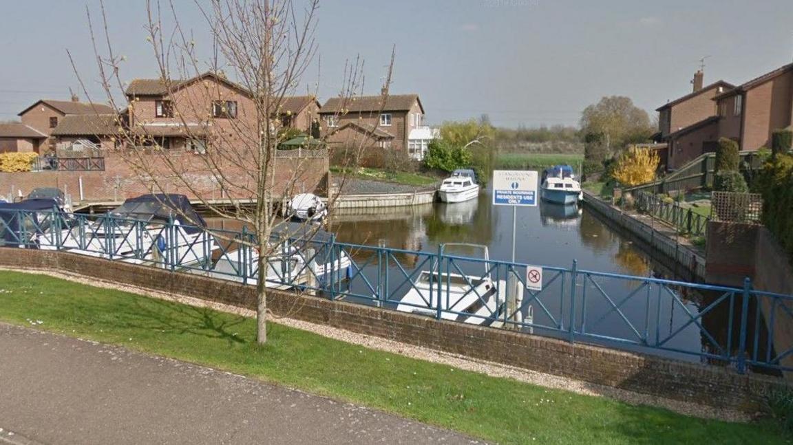 Riverside Mead, Peterborough, showing a piece of water, a bridge, boats on the water and houses either side of the river. There is a tree by the bridge.