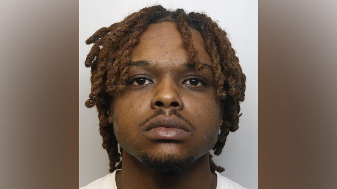 A police custody photograph showing a black man with dreadlocks and a small moustache, staring blankly forward