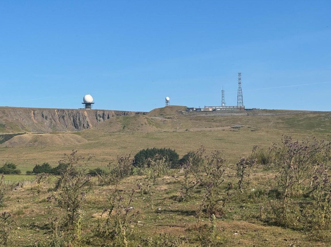 The circular structure and a similar one are in the distance. Fields and a hill dominate the foreground and the radomes are on top of the hill along with other structures.