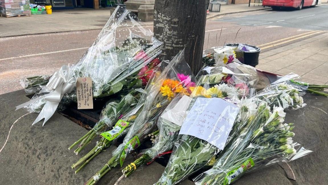 Flowers left around a tree in a street in Batley, West Yorkshire. There are several bunches of flowers, some with notes and ribbons on them.