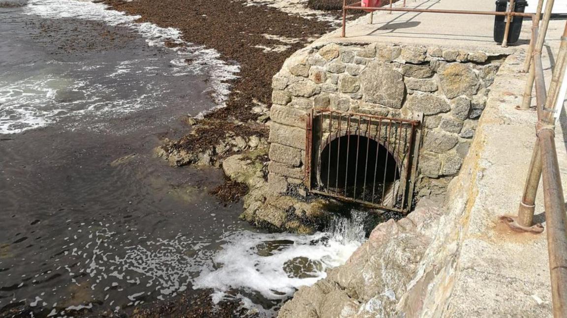 Storm overflow pipe at a beach in Cornwall. The pipe is coming from beneath a platform overlooking the sea. The hole is covered by a metal grate and liquid is pouring out of the pipe and into the sea.