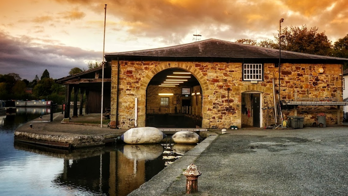 Scenic photo of part of the yard and canal featuring an old stone building next to the water with and orangey hue from the sun in the background