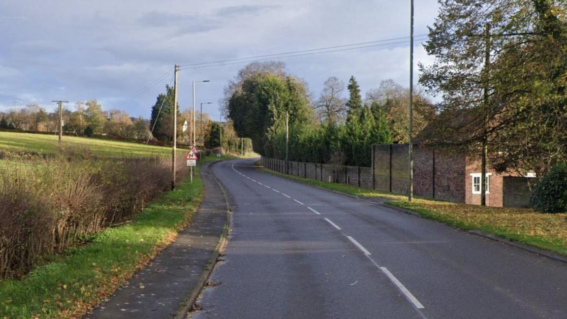 A stretch of road with hedgerows and a field to the left and a brown brick house and a long section of fencing and trees to the right