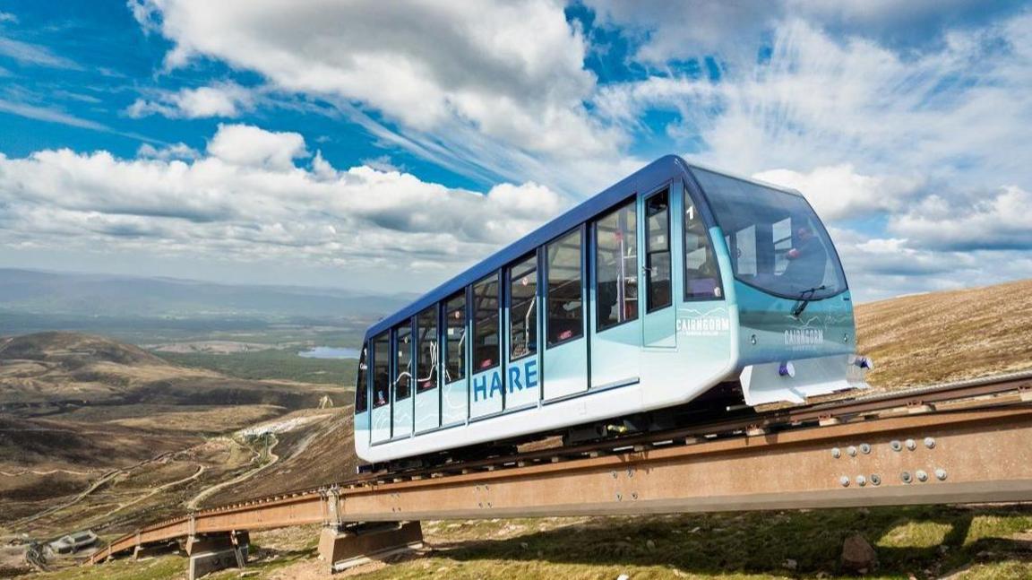 A tram-like carriage moves along a metal rail on a mountainside. The vehicle is blue and aqua coloured and has the word "hare" and "Cairngorm" on the exterior.