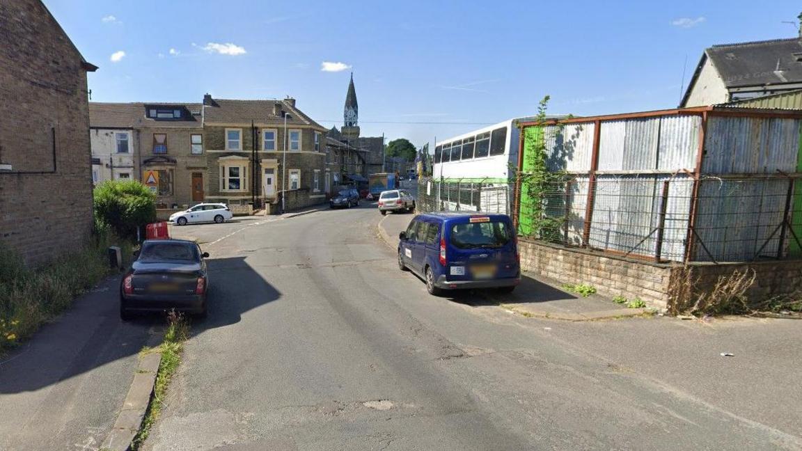 A picture showing a road with a purple car parked on the right hand side and another car parked on the left. The road also shows houses and industrial type premises. 