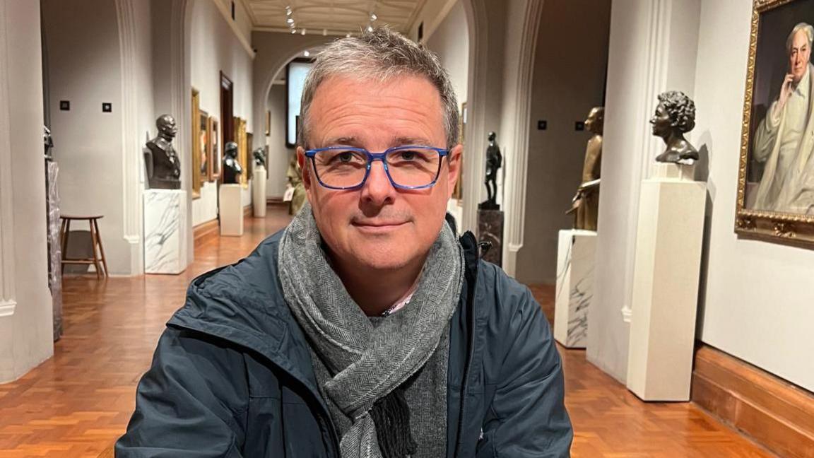 Niven smiles in a long art gallery. He wears blue framed glasses a hooded light jacket and a grey scarf. Behind him is a hallway lined with bronze statues and hanging framed artworks.