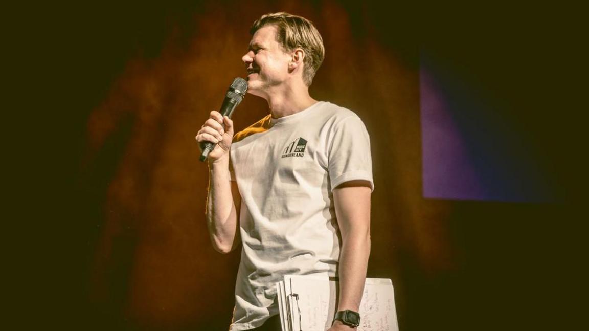 Side image of a man with short light brown hair wearing a beige T-shirt. He is holding up a microphone in his right hand, and has a bundle of papers under his left