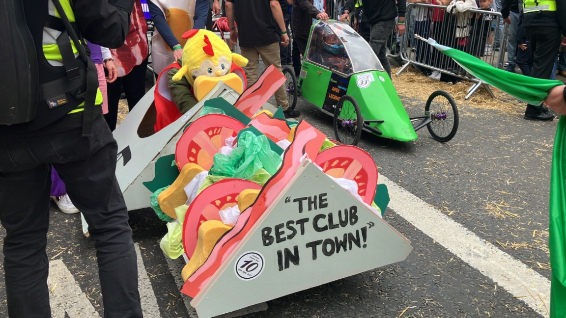 A kart which is shaped as a sandwich is on the left side of the road. It says THE BEST CLUB IN TOWN on the front. On the left is another kart which is green and has a window. 