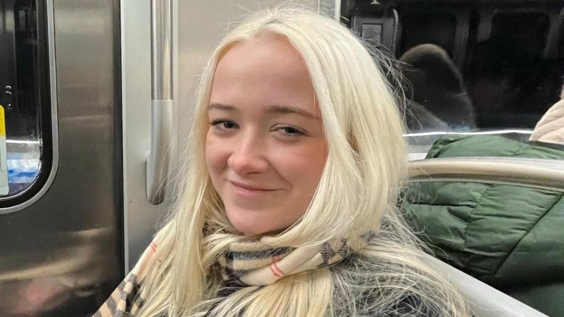 A woman sitting on a train and smiling at the camera. She has long blonde hair and is wearing a tartan patterned scarf. Behind her are metal train doors and a darkened window.