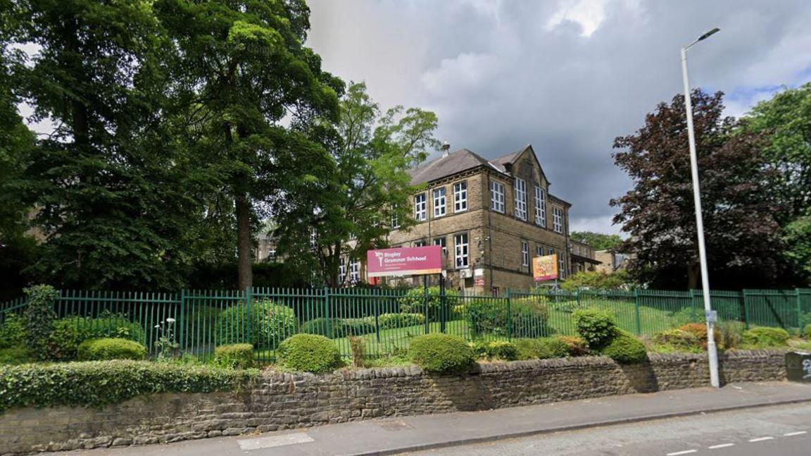 Bingley Grammar School which is a stone building set in grassed grounds with a stone wall and green metal fence around. There is a dark red sign which reads 'Bingley Grammar School'.