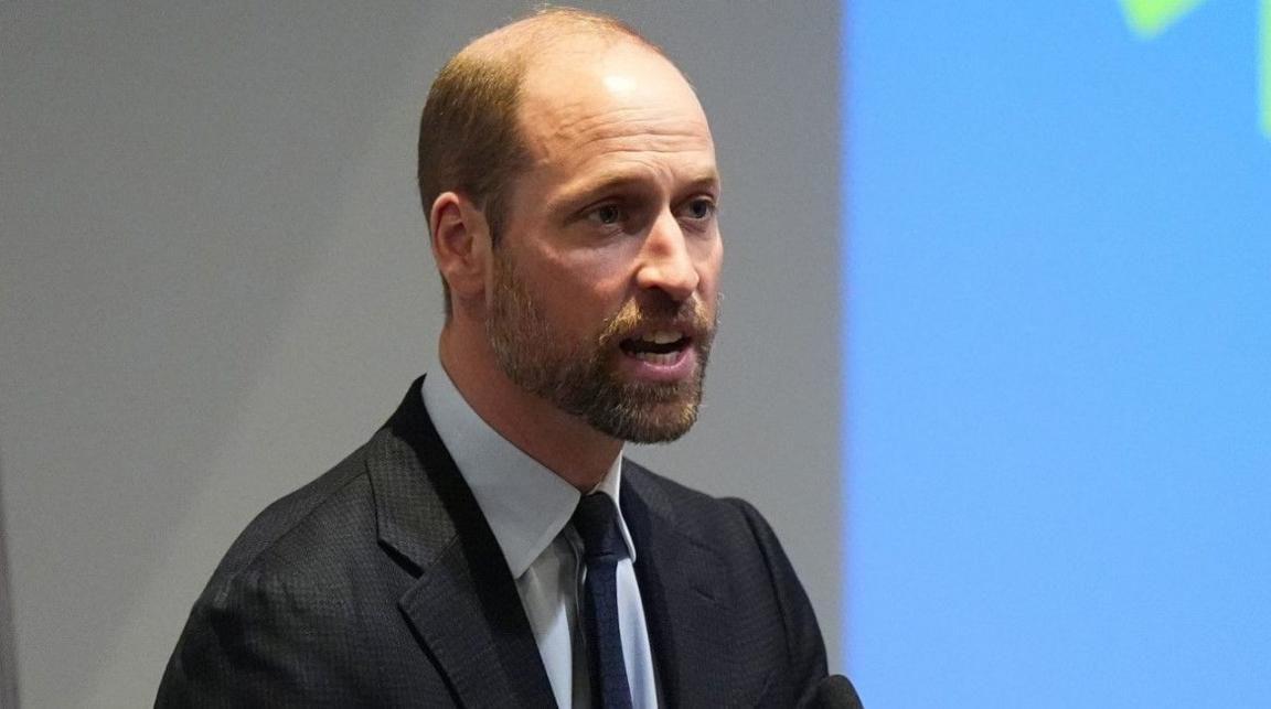 The Prince of Wales, with a beard, wearing a dark jacket and tie. He is standing near a blue background. 