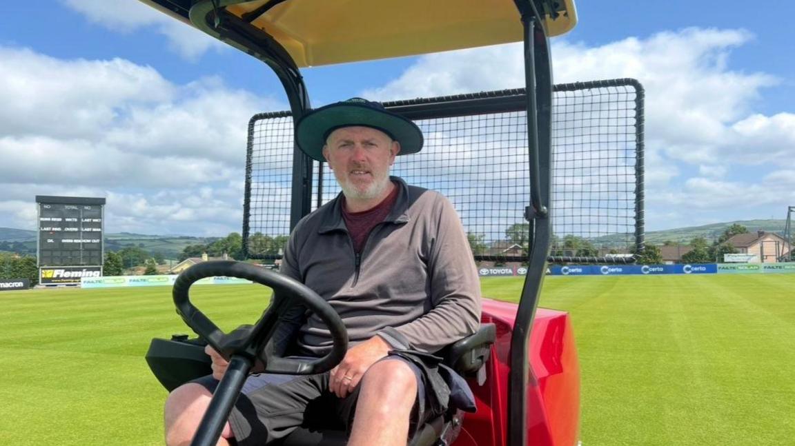 Trevor Hamilton is wearing a grey three quarter zip, wearing a green hat sitting on a red tractor. The tractor is on a green pitch.