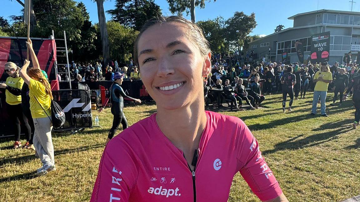 Megan smiles at the camera at the end of a triathlon. She is wearing a pink lyrica top and her hair is tied back. Behind her other triathletes are gathered and are wearing swim suits.