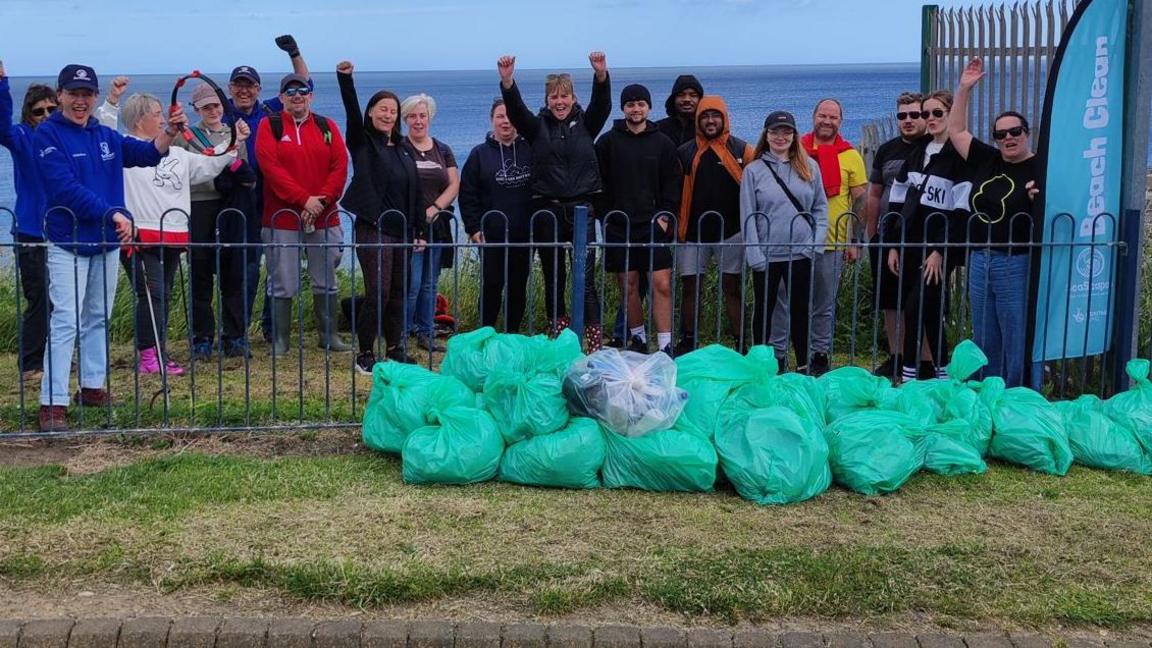 A group of volunteers standing in front of a beach with dozens of green bags of rubbish. Some are holding their arms in the air.