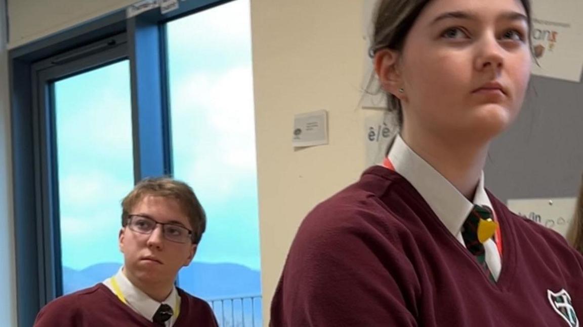 Richard and Marianne - students who took GCSE Latin at Keswick School. They wear maroon-coloured uniform. Richard, who wears dark-rimmed rectangular glasses is sitting in front of a large window through which the tops of mountains can be seen. Marianne, whose long dark hair is tied back, sits in front of him with both looking towards the same thing.