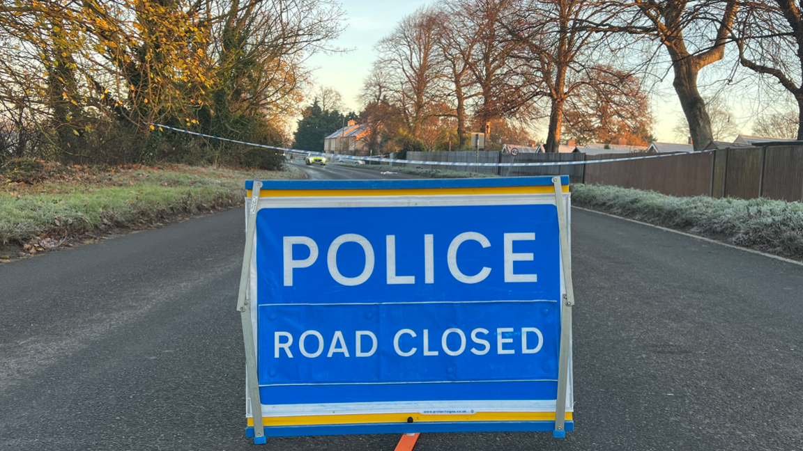 A blue sign in the centre of the road reads "Police Road Closed". Behind the sign is blue police tape, cordoning off part of Great North Road. 