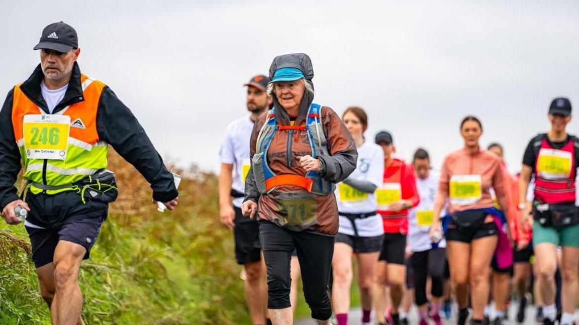 Walkers wearing taking on the challenge in brightly coloured sports wear with a grey sky in the background. The man at the front of the group has a grey beard and is wearing black orange and yellow top and black shorts and cap and has the walking number 246. An older woman wearing a who is wearing a blue, orange and grey backpack and a brown and grey coat with the hood up is slightly behind.