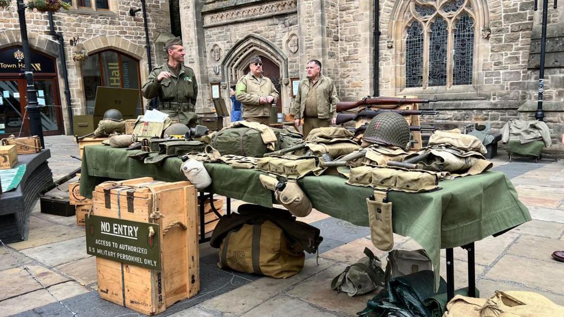 A table laid out with equipment from World War Two, including helmet, backpacks and water bottles. Three men in uniform of the time stand behind it.