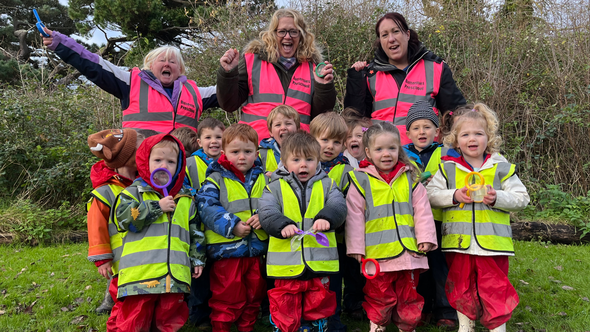 A group of young children from Butterflies pre-school kitted out in waterproofs and yellow high-vis jackets. Three staff members are behind them wearing their own coats and pink high-vis jackets. They are all cheering and holding mini plastic magnifying glasses. 