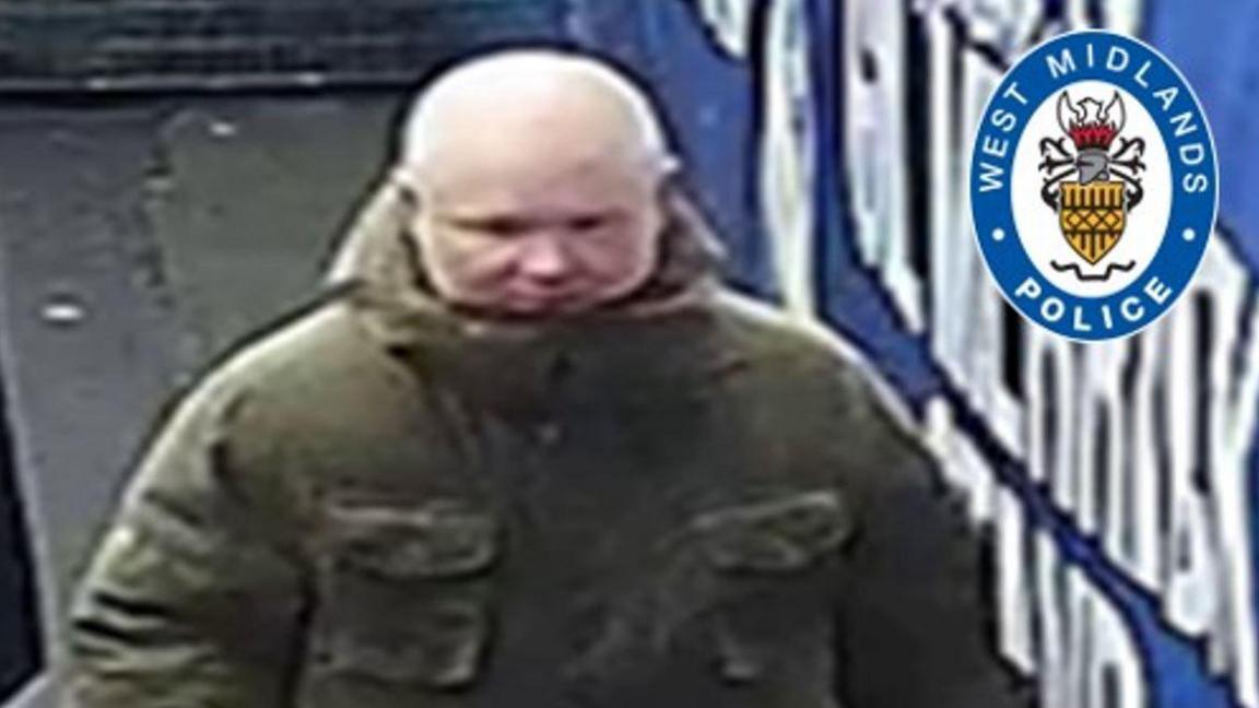 A bald man in a large green coat standing in the street alongside a large blue sign