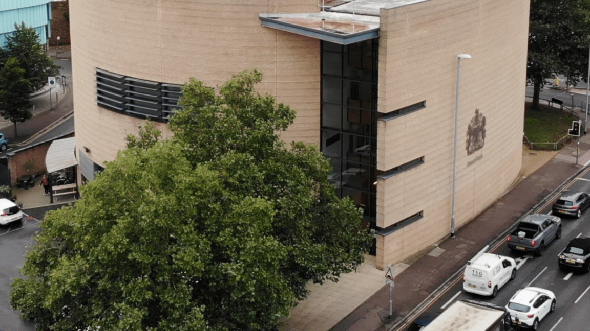 High angle of Cambridge Crown court building, built out of beige coloured stone  with a silver crown court symbol on the side. It is surrounded by trees, parking and a road full of traffic