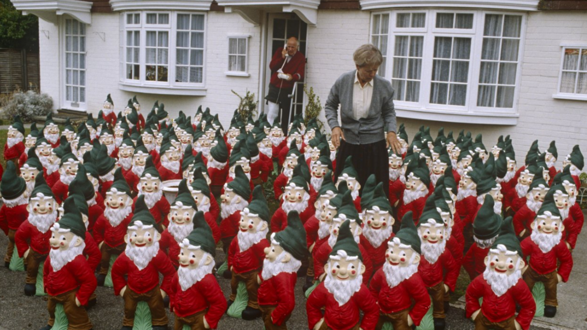 A scene from One Foot in the Grave with the front garden on Victor and Margaret's house covered in garden gnomes. Victor is standing at the front door on the phone and Margaret is in the middle of the gnomes.