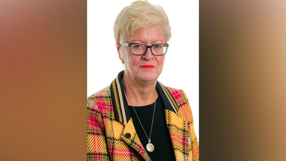 Chair of the CCRC Helen Pitcher wears a yellow and red jacket over a black top and smiles at camera. She has short blonde hair and glasses 