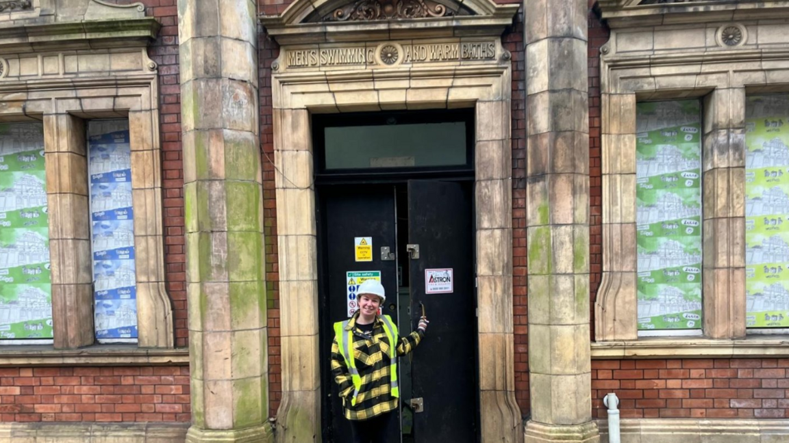 Emma Harvey stands in the doorway of the Jacob Wells Baths, wearing a hard hat and a florescent vest over a red and black top, with leggings. The Baths building is made of red brick with limestone bricks creating the doorway and windows. 